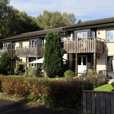 Ljus fastighet med två våningar på Evedalsgatan med träbalkonger och trädgård med staket och häck.