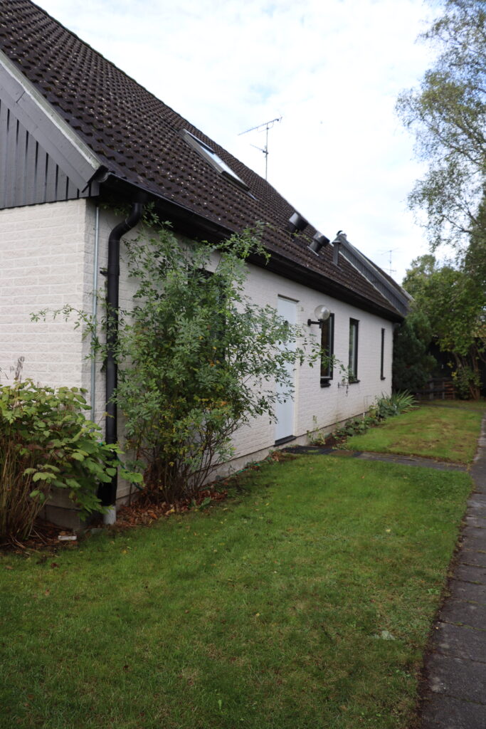 Grönalundsvägens fastighet från sidan, vit dörr och tre svarta fönster med grön gräsmatta utanför.