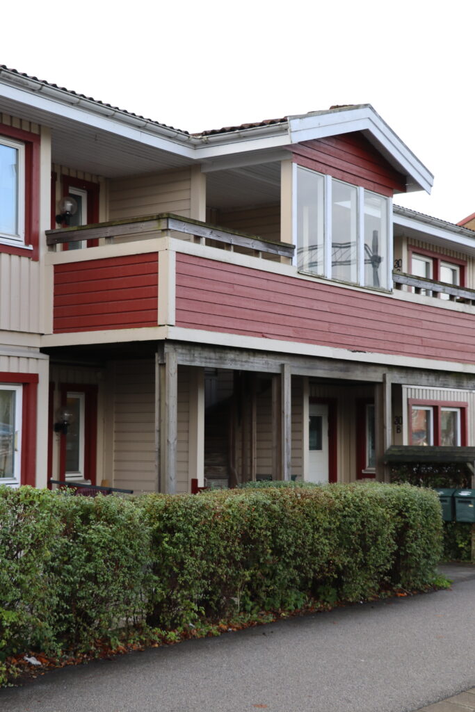 Närbild på Järnvägsgatan/östergatans fastighet med röd balkong, grön häck vid ingång.