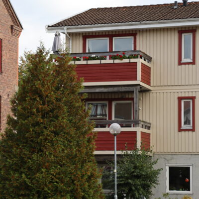 Gult trähus med två våningar, närbild på röda balkonger och hög gran till vänster.
