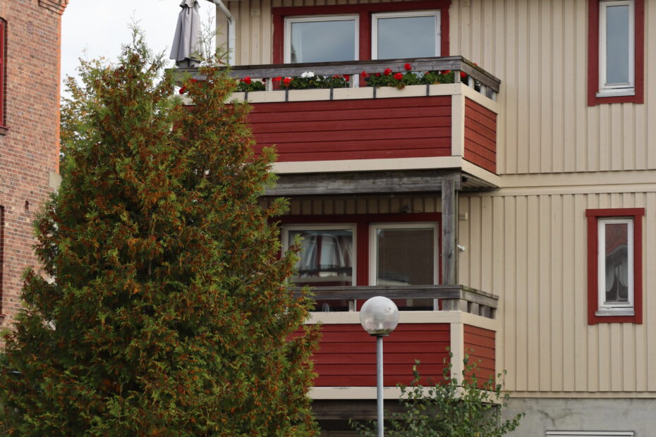 Gult trähus med två våningar, närbild på röda balkonger och hög gran till vänster.